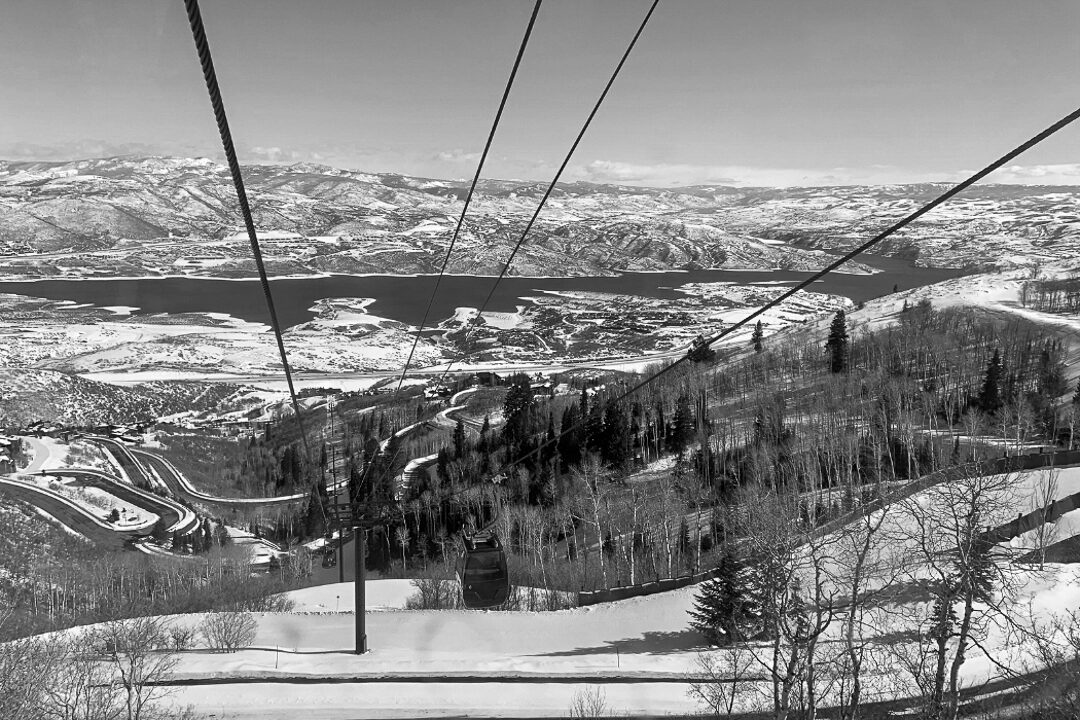 Looking out from a gondola over the Jordanelle Reservoir, with cables in the foreground and winding roads and ski trails weaving through snow‑dusted hills.